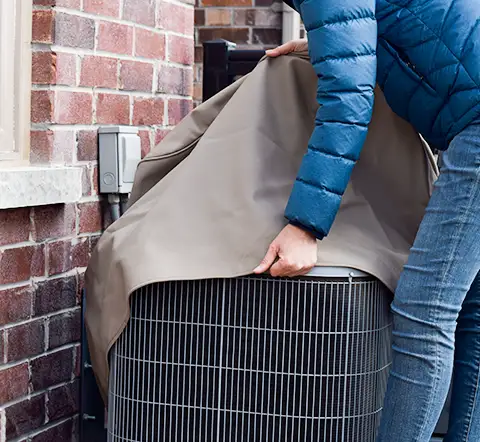 A man in a heavy jacket is putting a cover over his outdoor AC unit